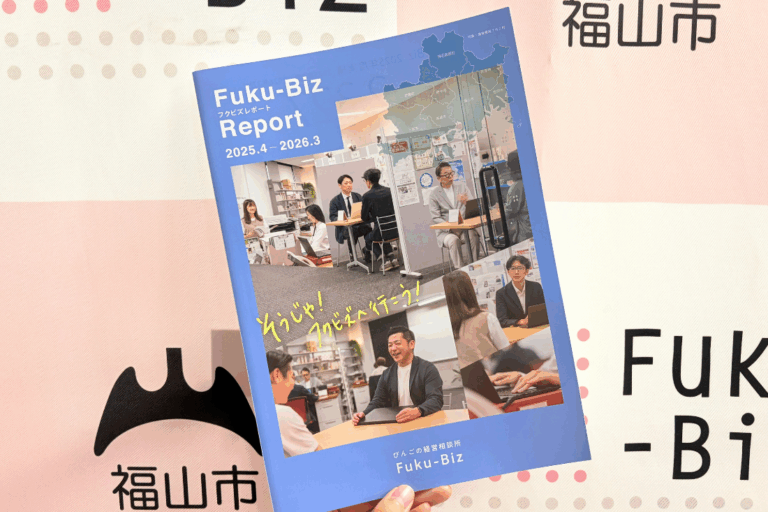 Cover of a blue Fuku-Biz Report magazine held in a hand, featuring office staff in discussions, a map graphic, and yellow handwritten text with the dates 2025.4–2026.3.