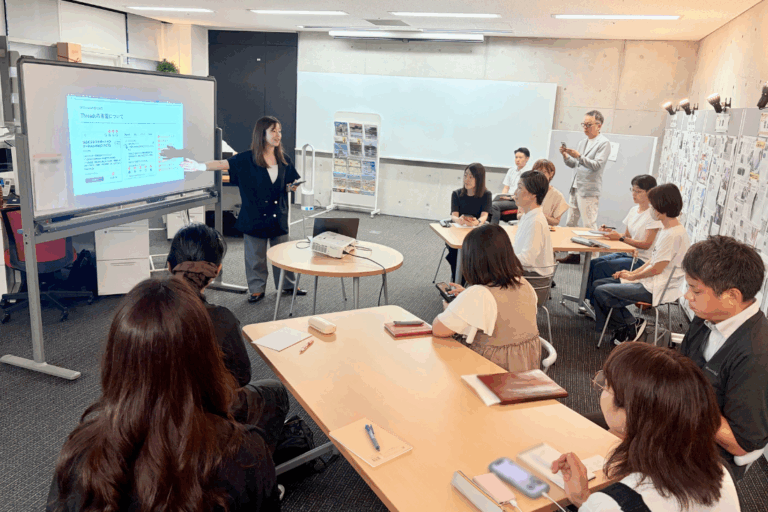 Trainer leads a workshop at a whiteboard screen, presenting to a semicircle of adults in a classroom. They take notes and watch attentively.