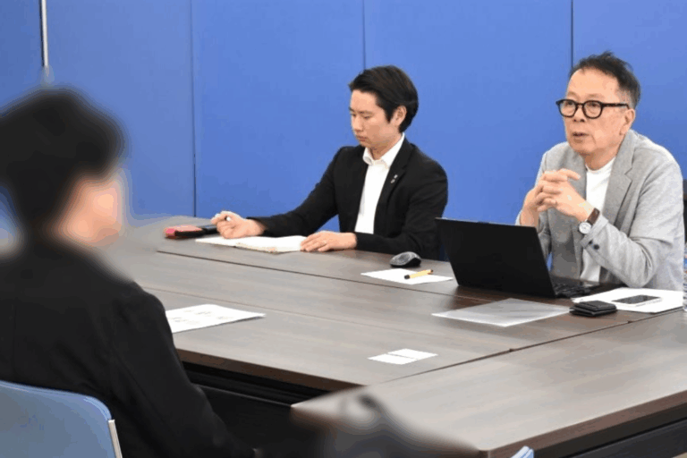 Two men sit at a conference table during a meeting: one in a black suit writing notes, the other in a gray blazer speaking with a laptop in front of him, while a blurred participant sits across from them in a blue-walled room.